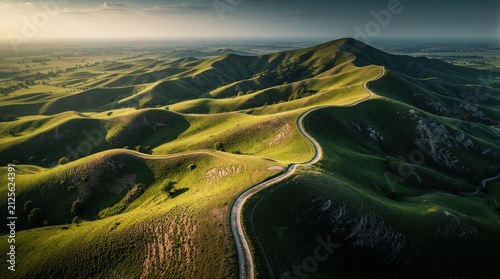 Aerial view of winding road through rolling green hills at sunset. Dramatic lighting creates high contrast shadows and highlights on textured landscape. Serene nature scene.