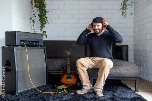 Obraz na plátně Hispanic musician sitting on couch to place on red headphones, electric guitar a