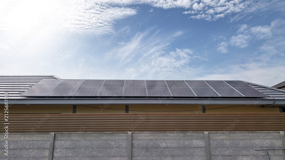 Fototapeta premium Residential rooftop solar panels installed on a house tile roof under a clear blue sky with white clouds. Clean renewable energy and sustainable living concept