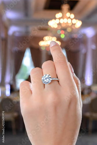 Close up of a luxury diamond engagement ring on female hand with blurred banquet hall background.
