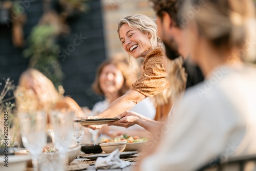 A laughing woman serves a plate of food at a crowded outdoor dinner table, capturing a natural hospitable moment for family restaurant marketing