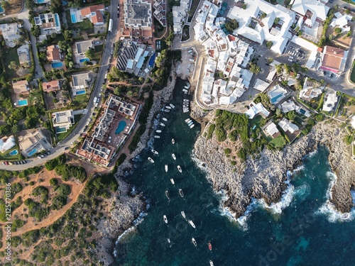 Aerial view of Binibequer Vell with white buildings and narrow streets along the rocky coastline and turquoise waters of the Mediterranean Sea in Binibequer Nou, Illes Balears, Spain.