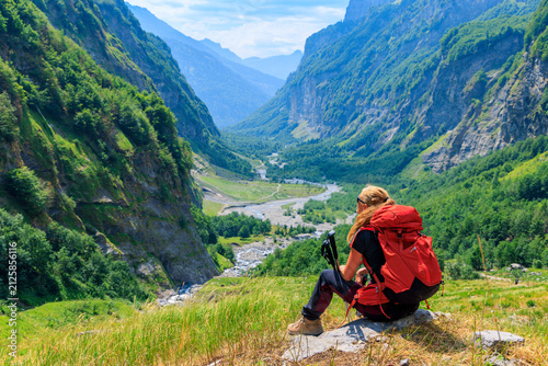 Female hiker with red backpack sitting on rock looking down at green mountain valley in Sixt Fer a Cheval, Alps in France, Chamonix