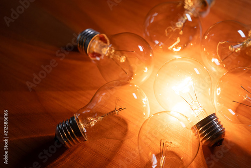 A group of transparent incandescent light bulbs lies on a wooden surface. One bright lamp glows among other turned off ones, expressing inspiration, creativity, and idea search
