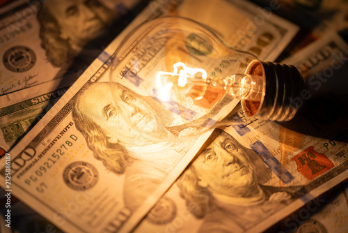 A glowing incandescent light bulb lies on a stack of one hundred dollar bills. A bright lamp illuminates Franklin's portrait on money, symbolizing success, wealth, and idea monetization