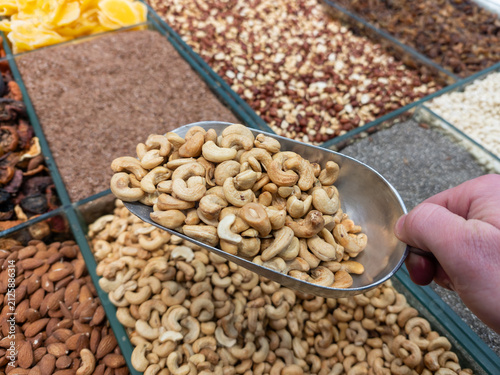 Metal scoop with cashew nuts in a store. A hand holds a scoop filled with cashew nuts against a display of assorted nuts, symbolizing choice and product quality
