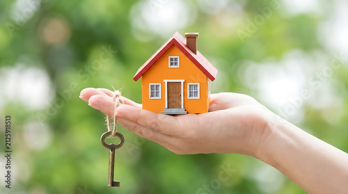 A hand holding a small orange toy house model with a metal key attached, symbolizing real estate, home ownership, and property investment
