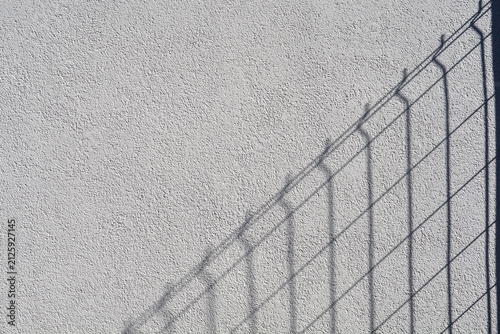 Фотография A diagonal shadow of a wire mesh fence is cast on a bright white textured stucco
