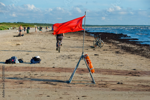 Red warning flag and lifebuoy on a windy sandy beach with people and bicycles in background.