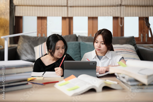 Two Asian teenage girls concentrate on online class, remote learning at home using tablet, writing and reading textbooks, self-study for back to school, Homeschooling, university education lifestyle.