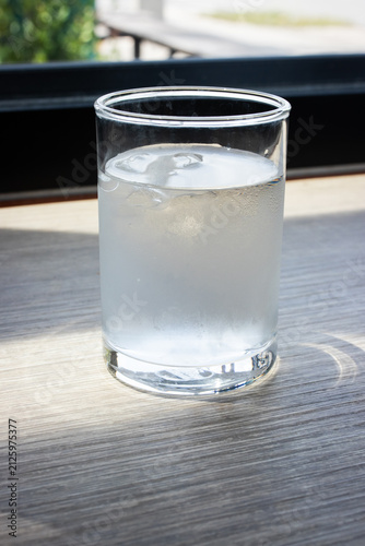 Glass of cold water with ice cubes under natural sunlight casting soft shadow on wooden table background.