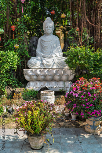 White stone Buddha statue sitting in lotus position in the courtyard of the Phap Bao Pagoda (Chua Phap Bao) surrounded by hanging lanterns and flowers in Hoi An, Vietnam.