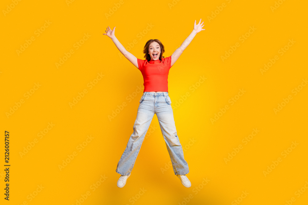 Obraz premium Happy young woman jumping with arms raised on bright yellow background expressing excitement and energy in a casual red top and wide-leg jeans