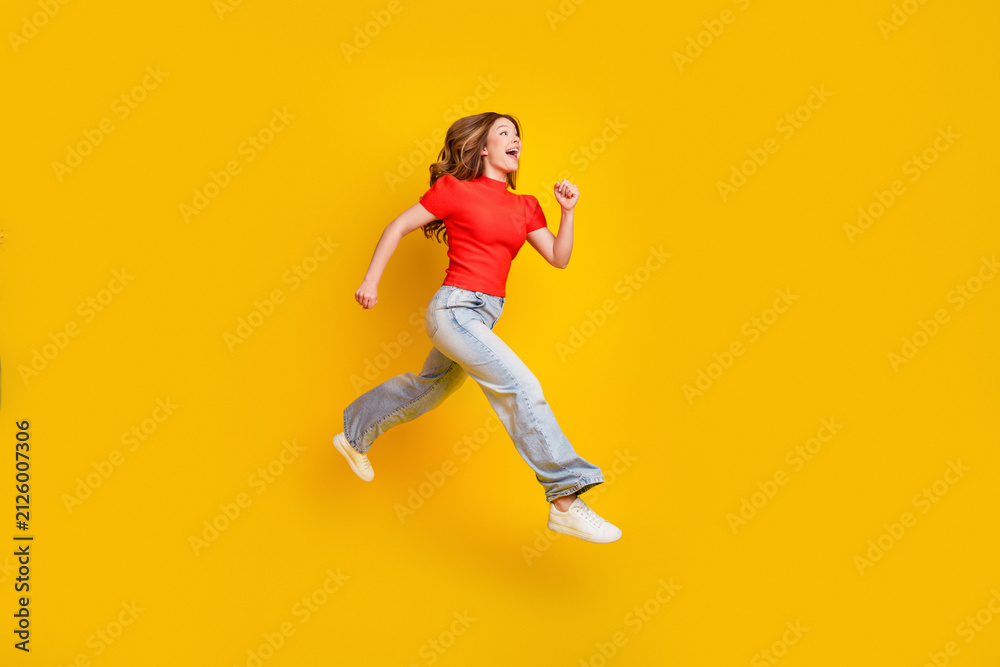 Fototapeta premium Young woman jumping joyfully against a yellow background in casual red shirt and jeans, energetic lifestyle and freedom expression