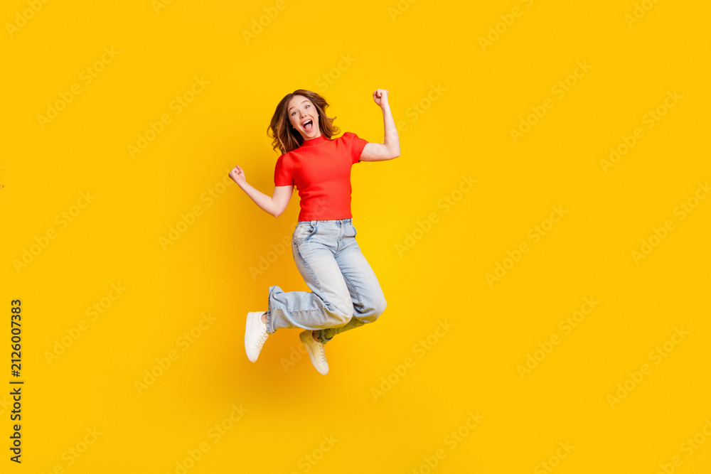 Fototapeta premium Happy young woman jumping in casual jeans and red top against bright yellow background celebrating joy and energy