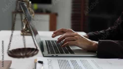 Lawyer typing on a laptop with a contract and scales of justice on the desk, female attorney working in a law firm office preparing legal documents for court