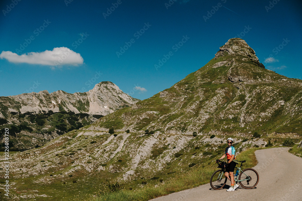 Fototapeta premium Female cyclist with gravel bike stopping on the mountain road at Sedlo Pass in Durmitor National Park, Montenegro. Stunning alpine scenery surrounds the scenic cycling route