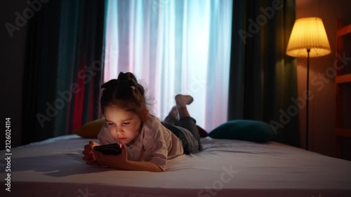 young girl is lying on the bed in the evening, holding a smartphone.