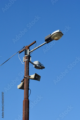 Bild auf Leinwand A low angle shot of a rusty metal utility pole with street lamps and public address horn speakers under a clear blue sky