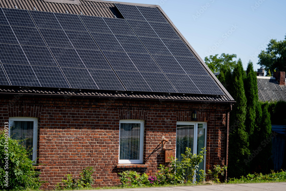 Fototapeta premium Solar panels installed on brick house roof with greenery and blue sky in background