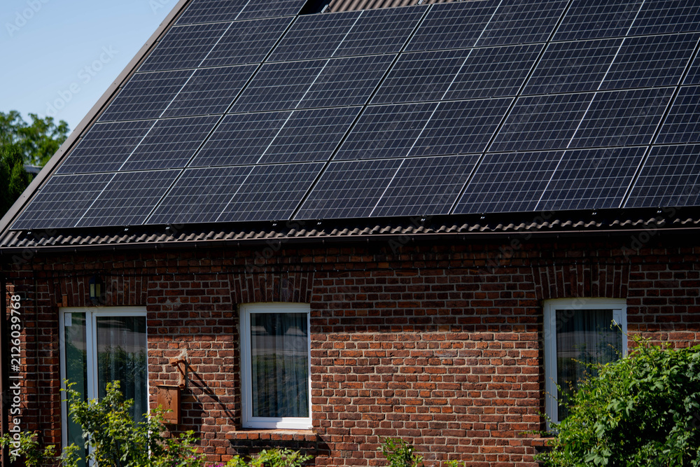 Fototapeta premium Solar panels installed on brick house roof with greenery surrounding the property