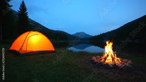 Campfire with tent beside lake at dusk.