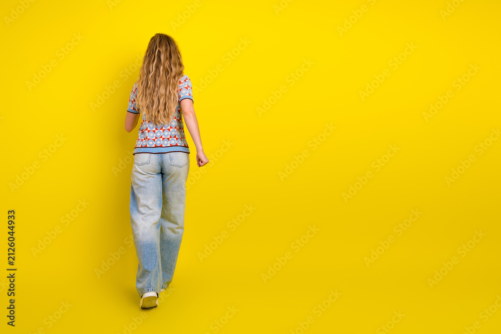 Fototapeta premium Young woman walking away on a bright yellow background in casual jeans and a patterned top, minimalist studio portrait conveying movement and carefree mood