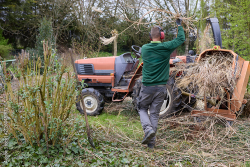 Fotografie Man operating a wood chipper, feeding dry branches into the machine attached to a tractor for garden clean-up