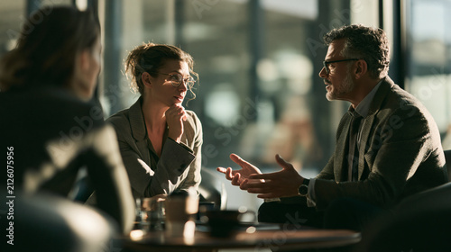 Business Meeting in Sunlight: Three professionals engage in a thoughtful discussion during a sunlit business meeting, fostering collaboration and strategic planning.