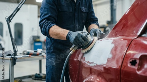 Auto mechanic using sander to polish car body in workshop. Repairman sanding metal surface of vehicle before painting. Professional automotive bodywork maintenance and restoration service.
