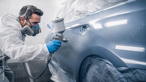 Painter using spray gun to paint car body in garage. Professional auto repairman in protective suit and respirator. Automotive bodywork service and vehicle restoration process.