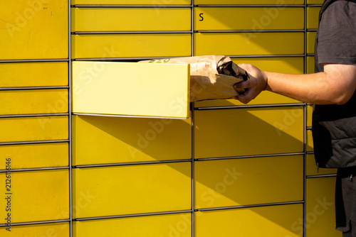 Person retrieving package from yellow parcel locker in urban setting during daylight hours
