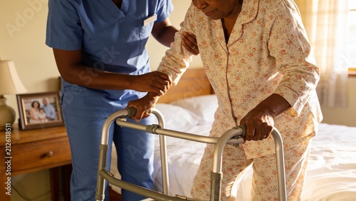 Senior woman using walker with caregiver assistance while getting out of bed in cozy bedroom.
