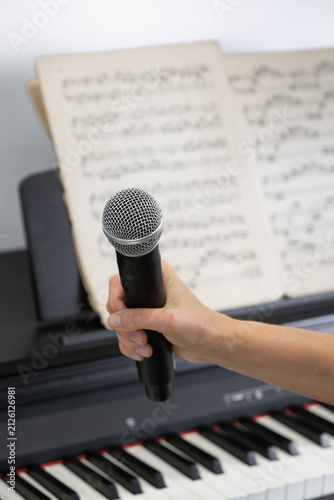 Child Holding Microphone During Singing Lesson with Piano and Sheet Music. Close-up of a child's hand holding a handheld microphone in front of sheet music on a digital piano.