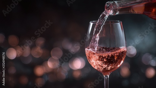 A glass of rose wine being poured from a bottle against a dark background with festive colorful bokeh lights for celebration