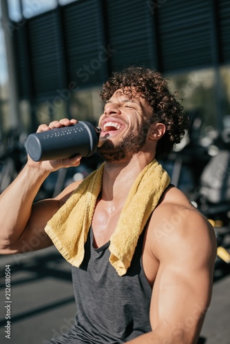 Smiling muscular man laughing while drinking protein shake at gym with yellow towel for fitness poster and lifestyle advertisement