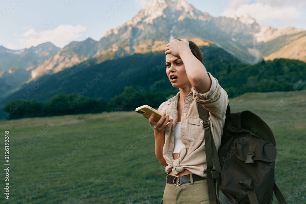 Obraz premium A young woman with a backpack stands in a mountain field holding a smartphone and looks confused while touching her head. Concept of outdoor lifestyle and navigation challenges.