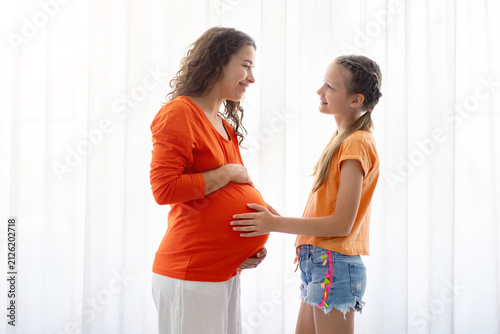 фотографія Pregnant mother and young daughter standing by bright window, smiling and bondin