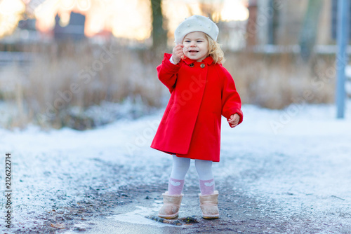 Outdoor winter portrait of little cute toddler girl in red coat and white fashion hat barret. Healthy happy baby child walking in the park on cold day with snow and snowfall. Stylish clothes for kids.