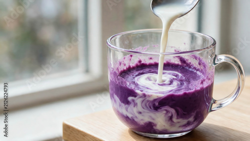 Close-up of cream swirling into a vibrant purple ube latte in a clear glass mug by a bright window, creating a smooth marbled pattern.