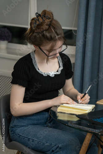 Focused young female student wearing glasses is taking notes in notebook while learning online using laptop, creating comfortable and productive learning space at home