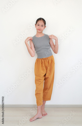 Pretty young Asian girl in ribbed gray top and rolled mustard yellow pants smiles while playfully lifting the hem of her shirt, posing bare feet on a wooden floor on white wall.