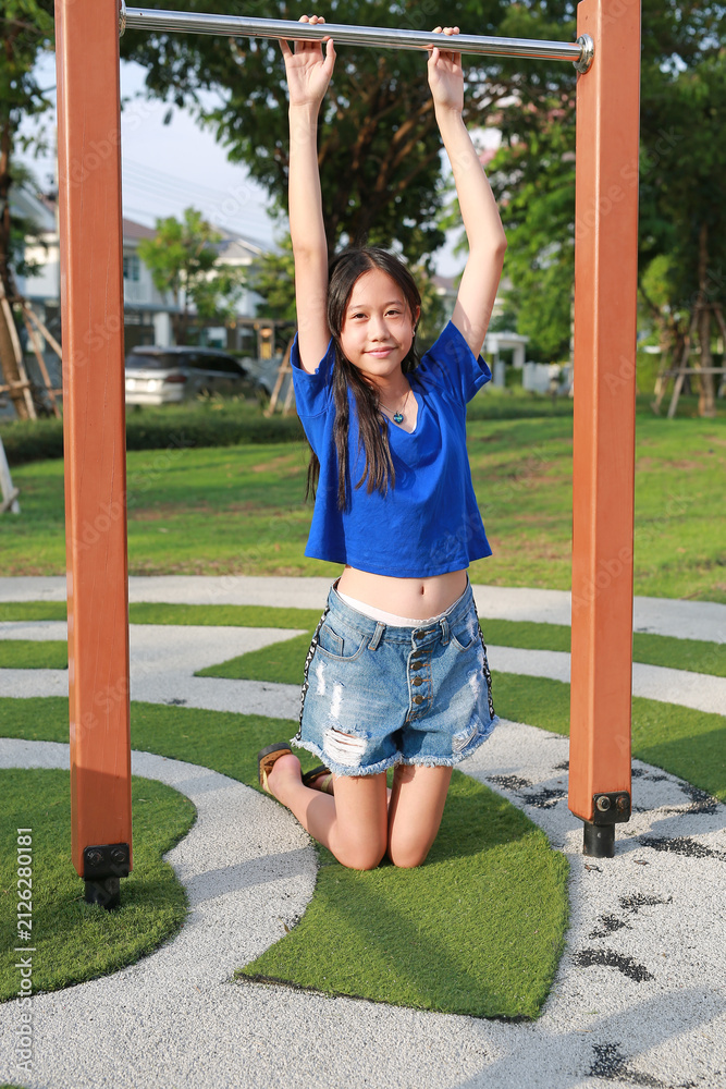 Obraz premium Cheerful young Asian girl holding onto the horizontal bar, enjoying outdoor playtime.