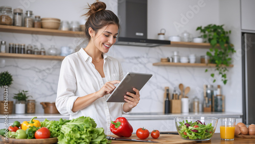 Smiling young woman using a tablet in a modern kitchen. Female preparing a healthy meal with fresh vegetables and reading an online recipe. Diet and nutrition concept