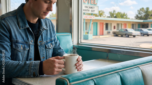 Man sitting in a retro diner holding a hot cup of coffee. View through the window of a vintage roadside motel. Concept of travel, nostalgia, and solitude during a road trip adventure.