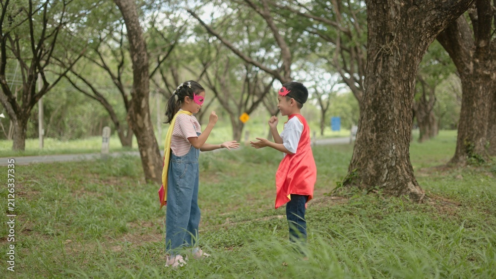 Fototapeta premium An Asian boy and girl wearing superhero capes doing a high five to celebrate successful teamwork while playing active games together in a green park area. area area area area