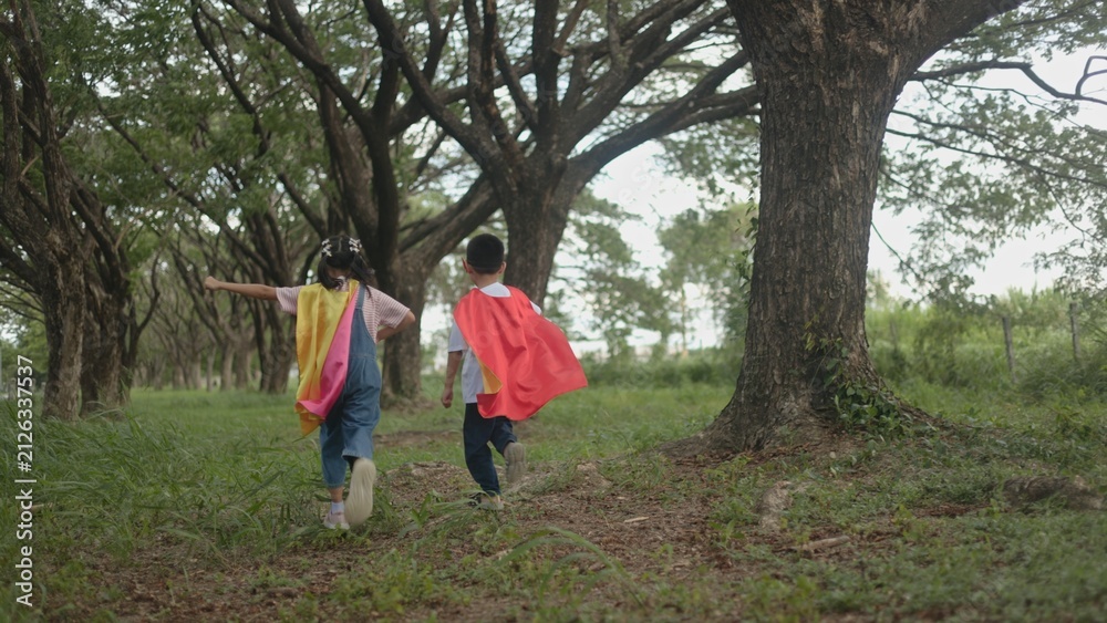 Fototapeta premium Two joyful Asian kids wearing superhero capes running together happily through a shaded park walkway during creative outdoor play time in a green park environment. area area