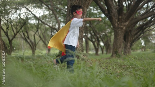 Konstfotografi An active Asian boy wearing a superhero cape running playfully across a green grass field under shaded trees in a peaceful public park environment during daytime