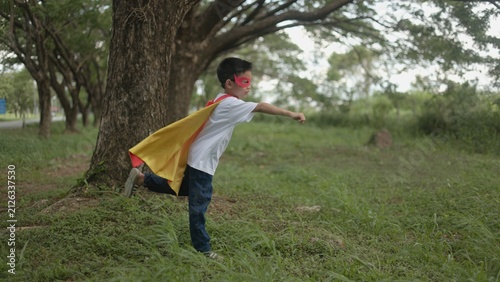 Canvastavla A brave Asian boy in a superhero costume running forward with his fist extended like a flying hero while playing active games in a green public park setting area