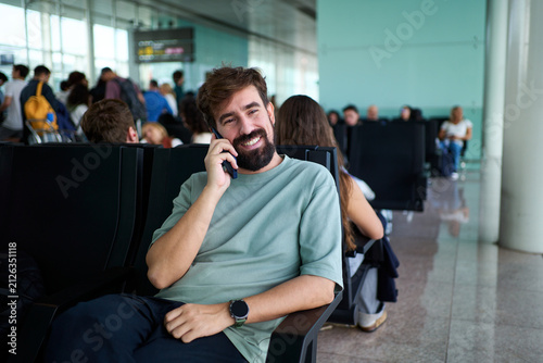 Smiling man sitting and enjoying a phone conversation while waiting for his flight at the airport departure area.